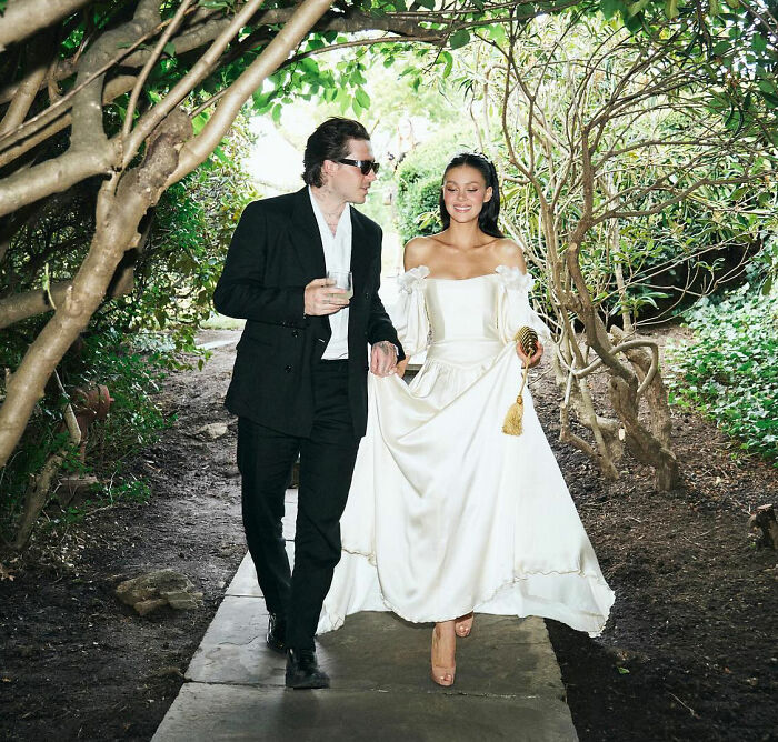 Brooklyn Beckham walking with a woman in a white dress under tree branches during an outdoor event. Brooklyn Beckham walking with a woman in a white dress under tree branches during an outdoor event.
