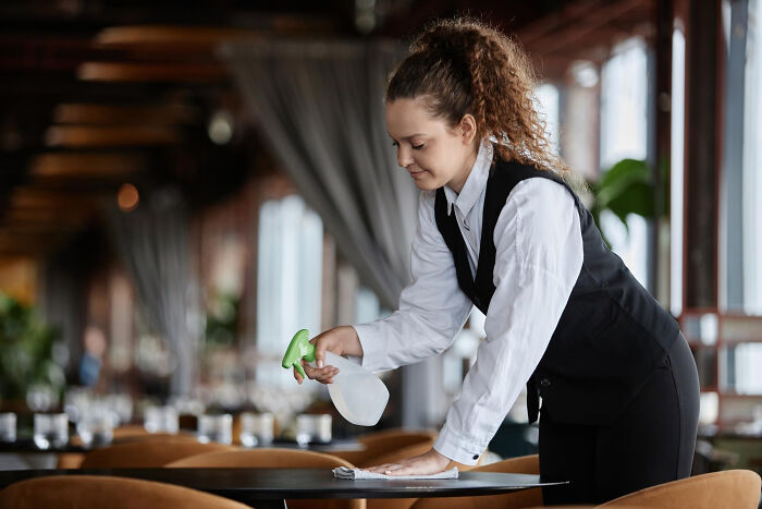 Young service industry worker cleaning table with a spray bottle and cloth in a restaurant during awkward moments at work