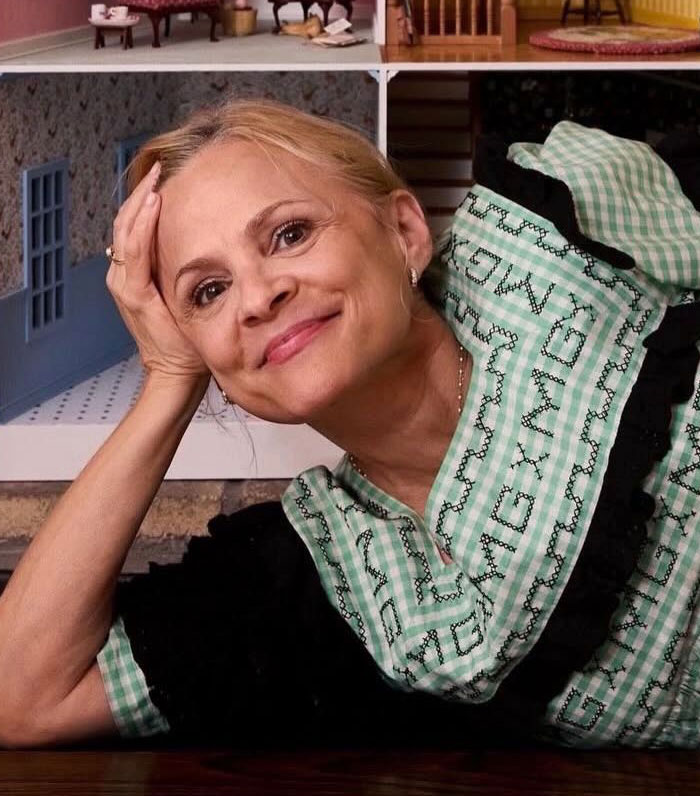 Amy Sedaris smiling and leaning on her hand, wearing a green patterned dress in a cozy indoor setting.