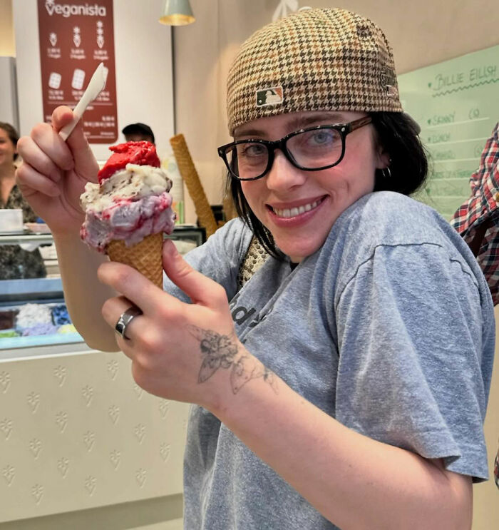 Smiling brunette in cap and glasses holding a triple-scoop ice cream cone inside a gelato shop Smiling brunette in cap and glasses holding a triple-scoop ice cream cone inside a gelato shop