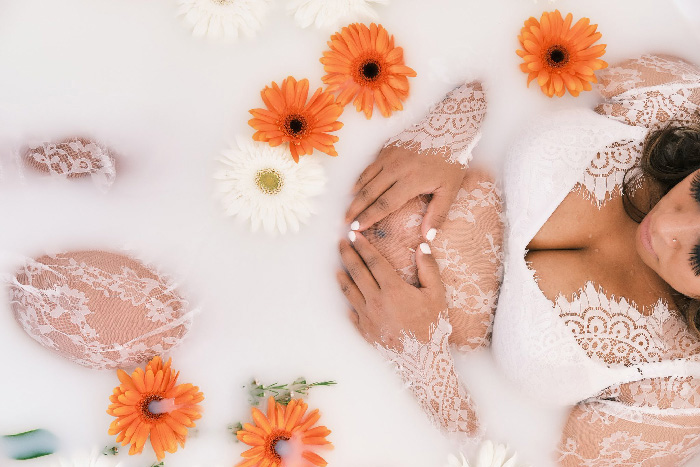 Pregnant woman in lace gown in milk bath with orange and white gerbera daisies, hands on belly, maternity photoshoot