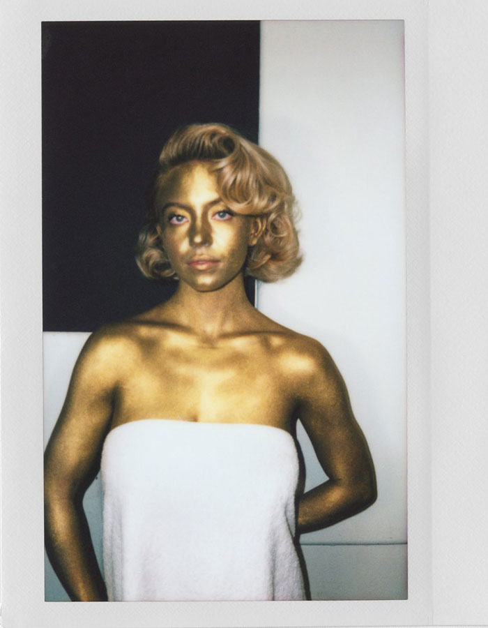 Gold-painted woman wrapped in towel with vintage curled hair, posing in a celebrity photoshoot against a black and white backdrop