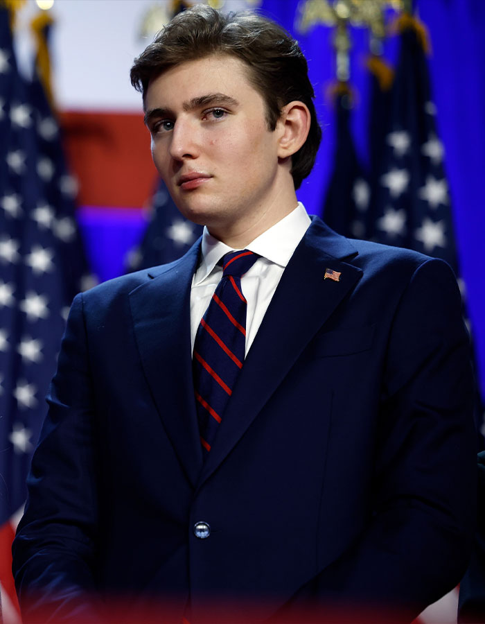 Barron Trump in a navy suit and striped tie, standing in front of American flags at a formal event. Barron Trump in a navy suit and striped tie, standing in front of American flags at a formal event.