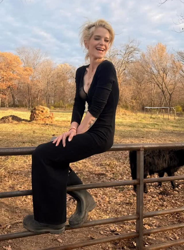 Kelly Osbourne sitting outdoors on a fence, smiling and wearing a black dress and boots in a rustic setting.
