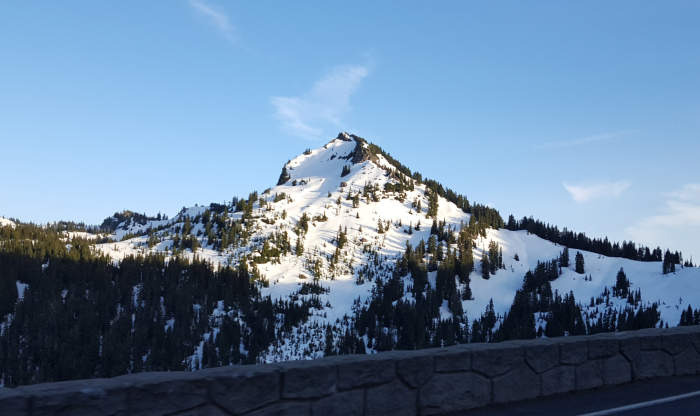 Snow-covered mountain with dense forest below under clear blue sky, illustrating haunted places in America’s eerie landscapes.