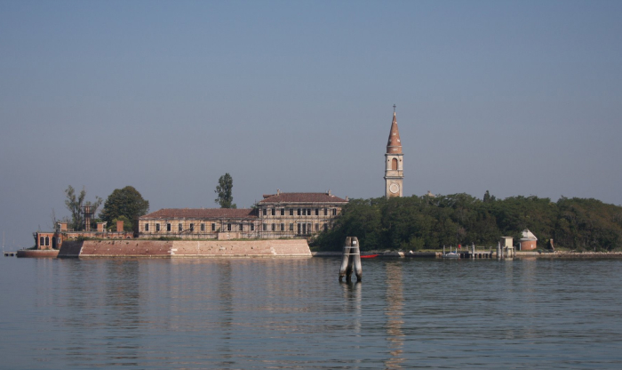A calm water view of a forbidden place on Earth with historic buildings and a tall clock tower surrounded by trees.