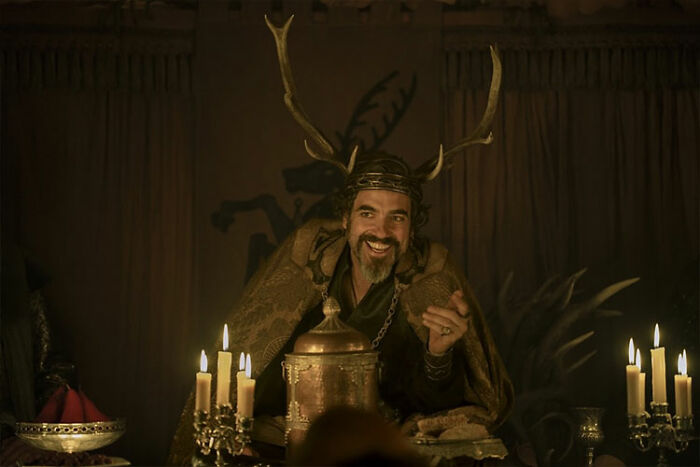Medieval character smiling with antler crown, surrounded by candles, in a scene from A Knight of the Seven Kingdoms ahead of season 1.