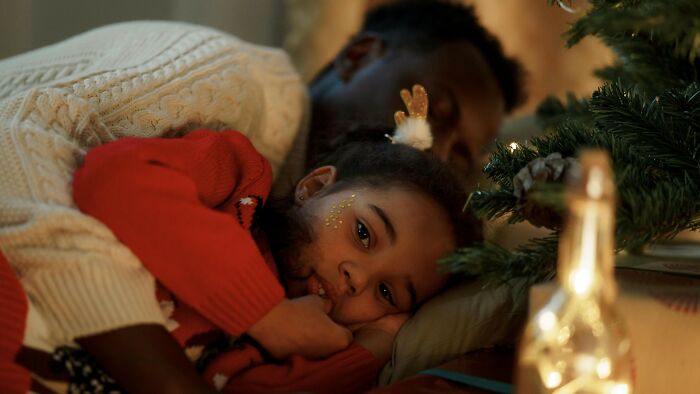 A child wearing reindeer antlers lying beside an adult near a decorated Christmas tree, reflecting on their choices.