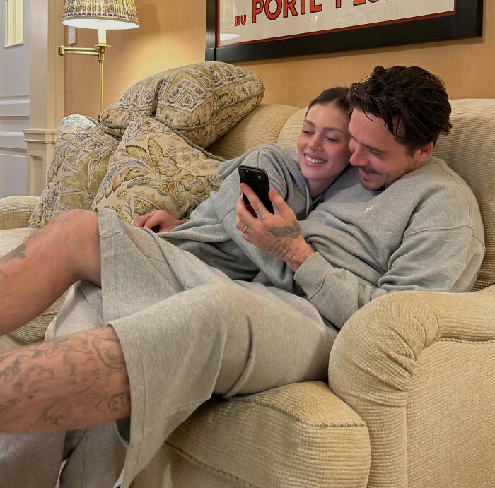 Brooklyn Beckham in casual wear, sitting on a couch with a woman, holding a phone, while family feud rumors swirl. Brooklyn Beckham in casual wear, sitting on a couch with a woman, holding a phone, while family feud rumors swirl.