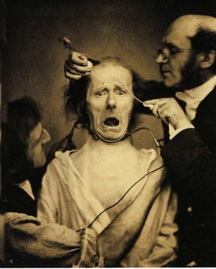 Old black and white creepy photo showing a distressed person being examined by two men in a vintage odd medical setting