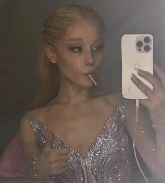 Ariana Grande taking a selfie with a smartphone, showing bizarre mannerisms and an unsettling expression. Ariana Grande taking a selfie with a smartphone, showing bizarre mannerisms and an unsettling expression.