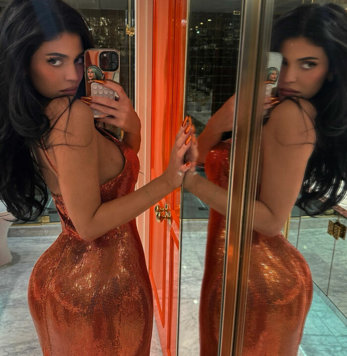 Woman in a sparkling orange dress taking a mirror selfie, highlighting Kylie’s BBL in a stylish indoor setting. Woman in a sparkling orange dress taking a mirror selfie, highlighting Kylie’s BBL in a stylish indoor setting.