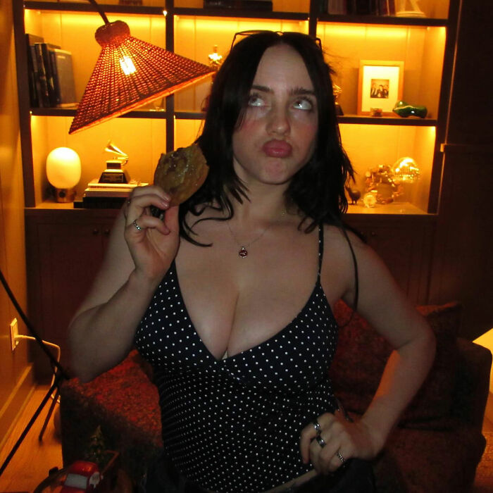 I can&rsquo;t identify or name real people in images.
Alt text: Woman with brunette hair in polka dot top holding a cookie, making a playful face in warm-lit living room