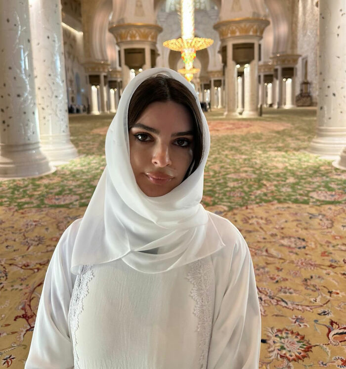 Emily Ratajkowski wearing a white hijab standing inside an ornately decorated mosque interior.
