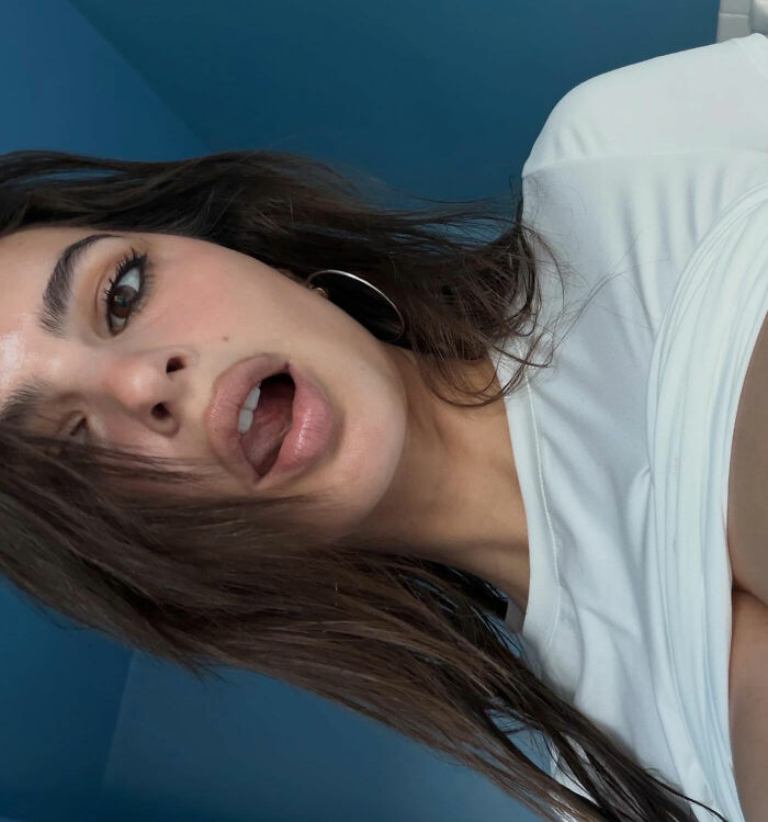 Emily Ratajkowski in a close-up selfie with tousled hair and hoop earrings against a blue background.