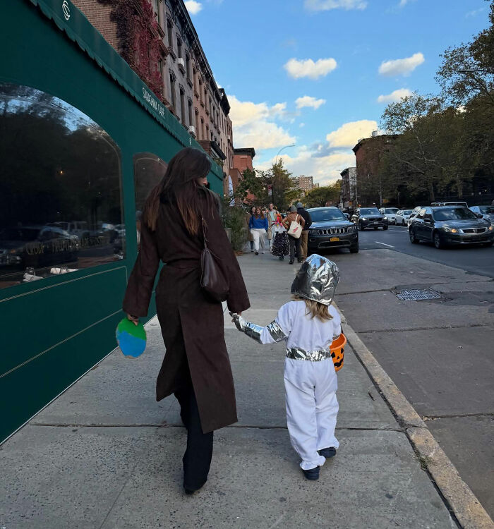 Woman walking on busy city sidewalk holding a child's hand dressed in a silver and white costume for Halloween.