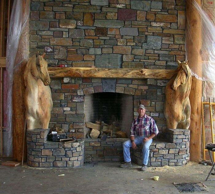 Man sitting by a stone fireplace with beautifully crafted woodworking projects featuring large horse head carvings on each side.