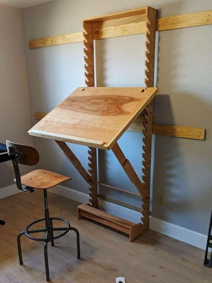 Adjustable wooden drafting table mounted on a wall next to a wooden and metal stool in a small room.