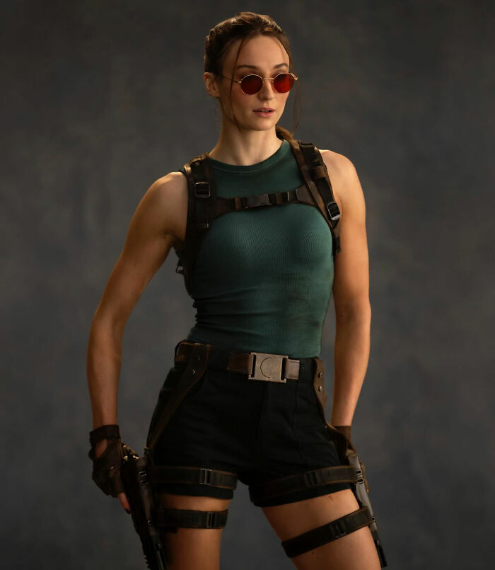 Sophie Turner in a ripped Lara Croft outfit with sunglasses and tactical gear, showcasing her Tomb Raider transformation.