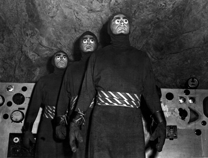 Three eerie figures in dark costumes with wide eyes stand in a vintage black and white odd and creepy scene.