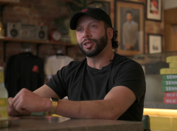 Pizzeria owner wearing black shirt and cap, sitting inside his restaurant, known for salty replies to harsh online reviews. Pizzeria owner wearing black shirt and cap, sitting inside his restaurant, known for salty replies to harsh online reviews.