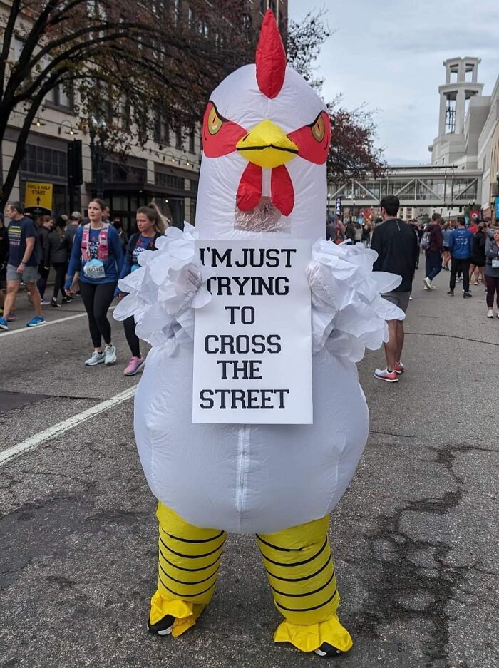 Person in a chicken costume holding a sign on a city street during a fun siblings-themed event with a crowd around.