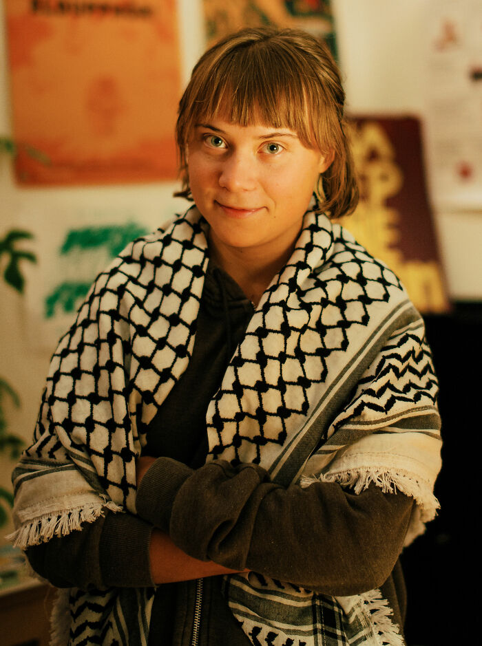 Greta Thunberg wearing a patterned shawl, smiling softly in an indoor setting with posters in the background.