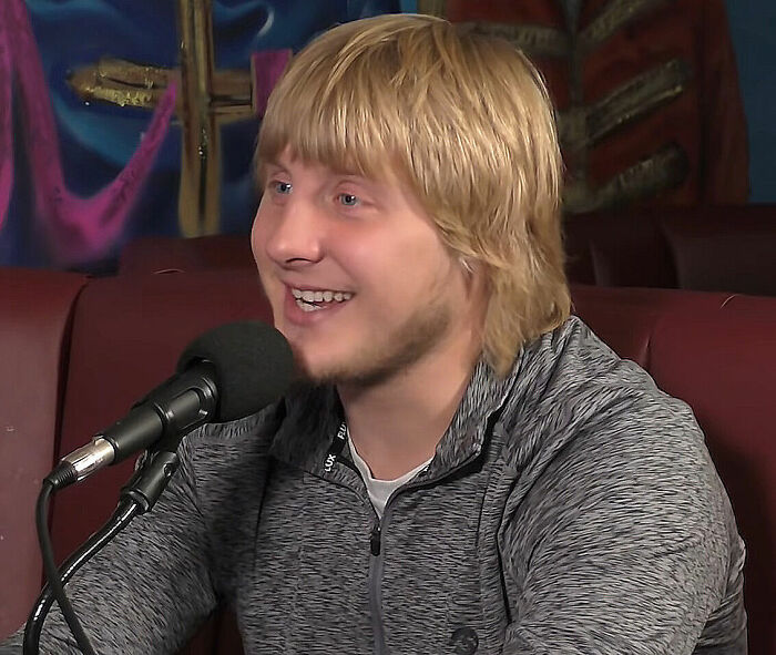 Paddy Pimblett speaking into a microphone during an interview, wearing a gray zip-up jacket indoors.