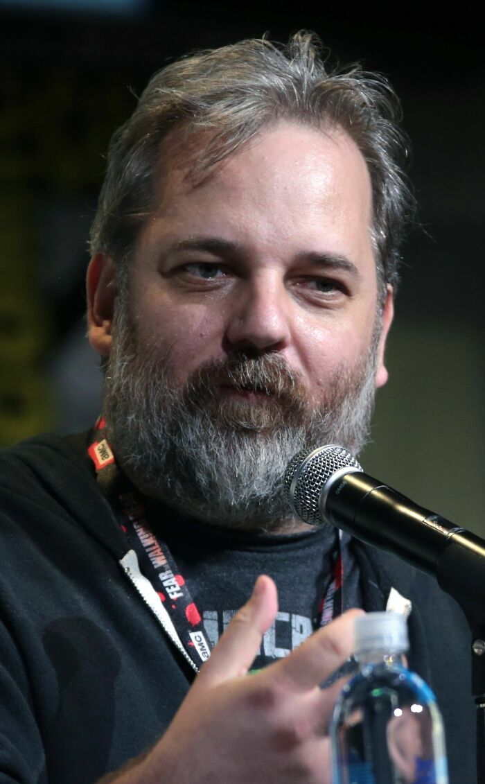Dan Harmon speaking into a microphone at an event, wearing a dark hoodie and a lanyard around his neck.