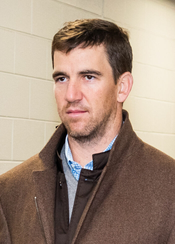 Eli Manning in a brown coat and blue shirt, looking thoughtful in an indoor hallway setting focusing on his career highlights.
