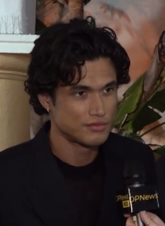 Charles Melton speaking in an interview, wearing a black jacket with a microphone labeled DPNews.