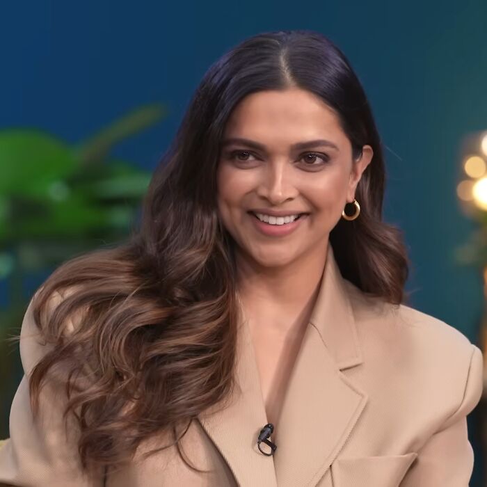 Deepika Padukone smiling with wavy hair, wearing a beige blazer and gold hoop earrings in an indoor setting.