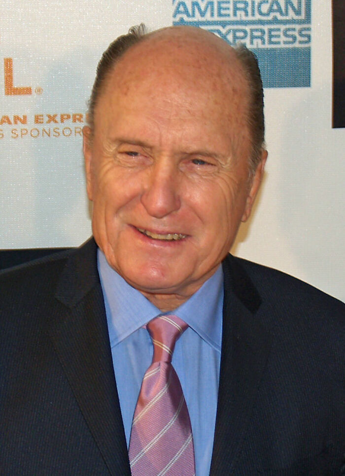 Robert Duvall smiling at an event, wearing a dark suit, blue shirt, and pink striped tie, highlighting his career.