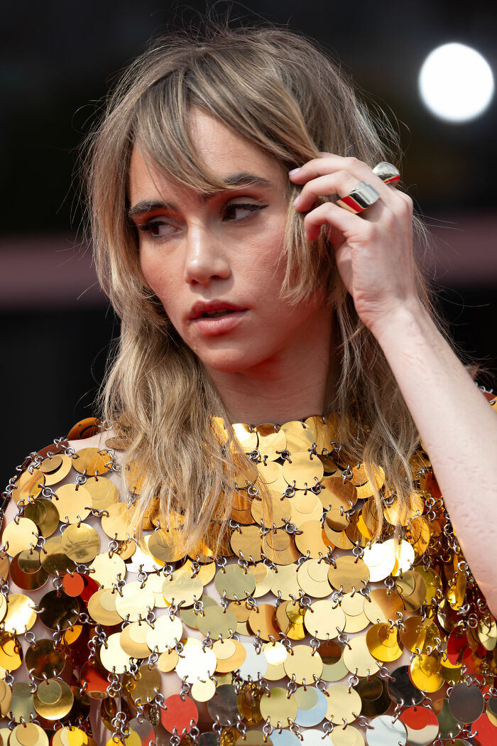 Suki Waterhouse wearing a shimmering gold sequin dress, touching her hair during a red carpet event.