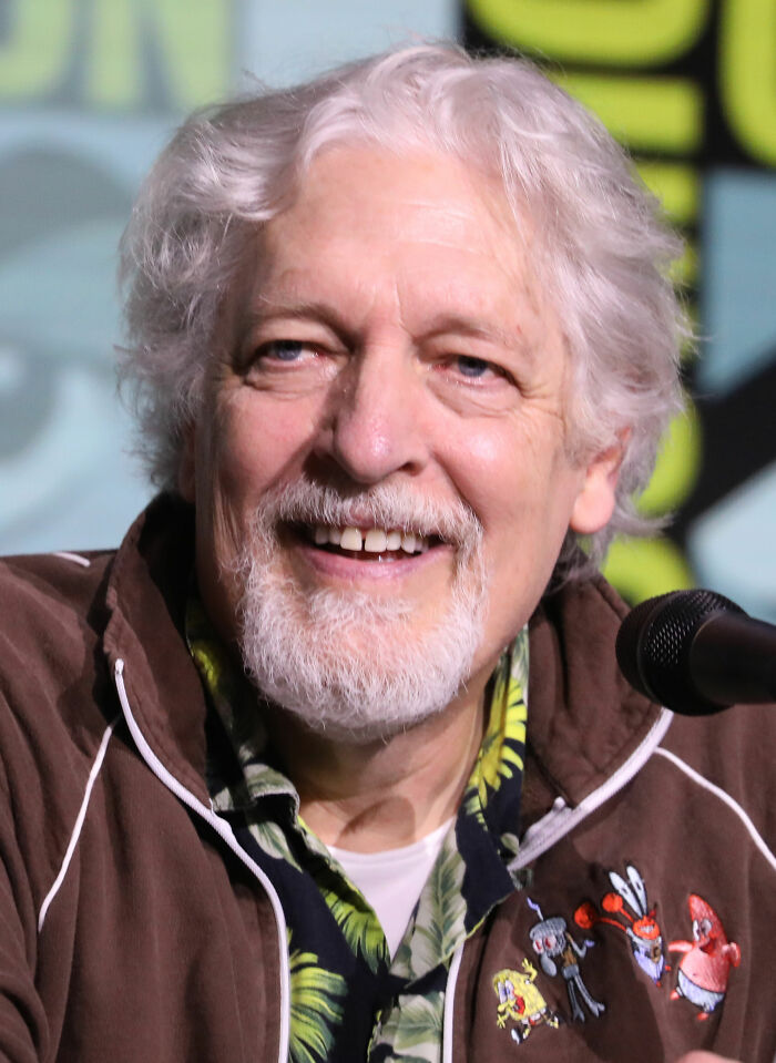 Clancy Brown speaking at a panel, wearing a brown jacket with animated character patches and a floral shirt.