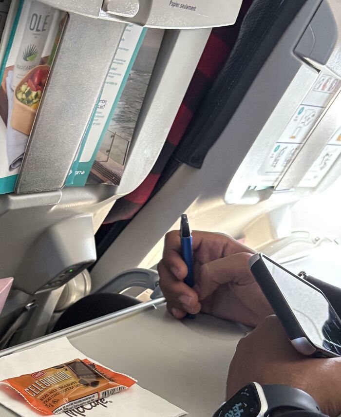 Passenger leaning over tray writing with pen, phone and snack visible in cramped airplane seat next-level jerks