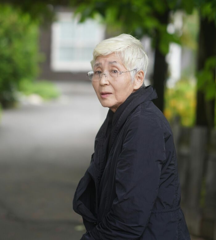 Older woman with short white hair and glasses outdoors, reflecting on family secrets in a quiet, leafy setting.