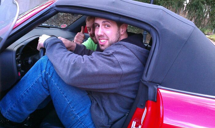 Two tall men struggling to fit comfortably inside a small red convertible car outdoors.