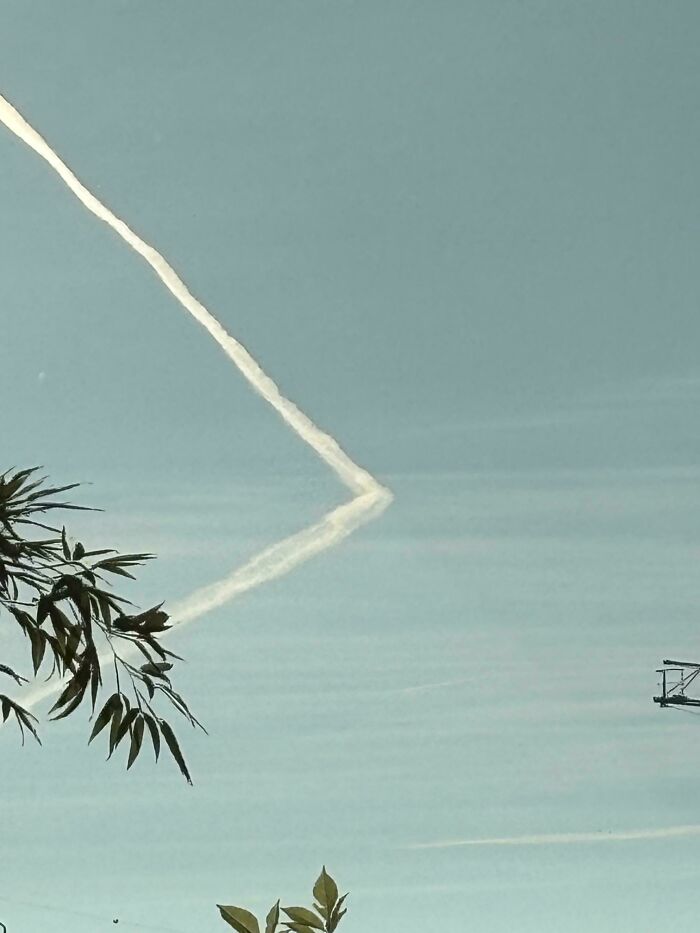 Contrail forming a zigzag in blue sky with leaves in foreground, evoking glitches in the matrix.