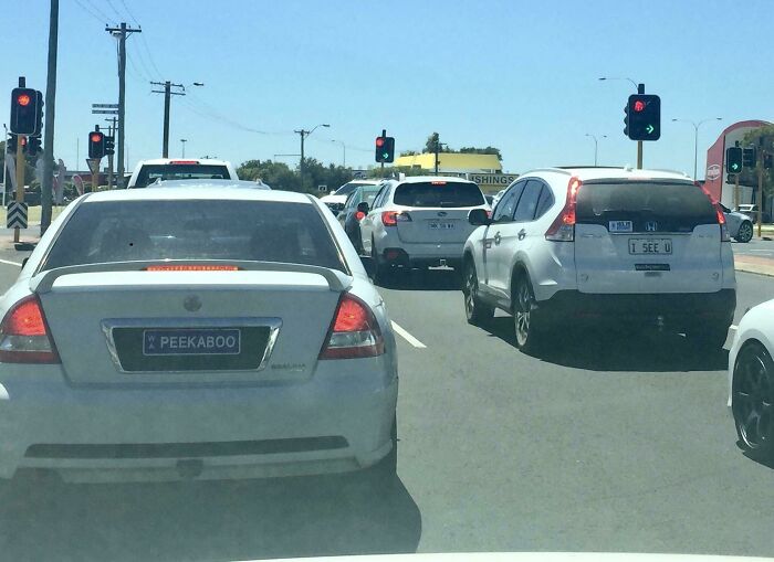 Cars stopped at an intersection on a sunny day, novelty plates reading PEEKABOO and I SEE U hinting at glitches in the matrix