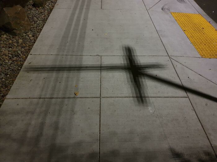 Sidewalk at night with a dark cross-like shadow and yellow tactile pad, evoking glitches in the matrix.