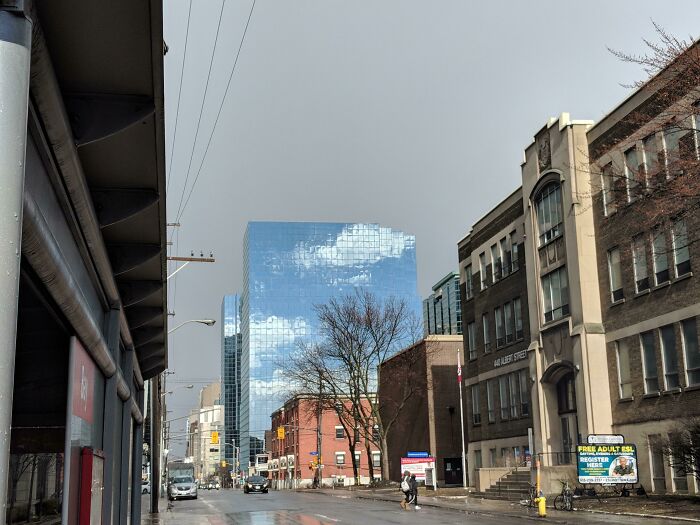 Urban street with reflective glass tower showing clouds, wet pavement and a pedestrian, evoking glitches in the matrix