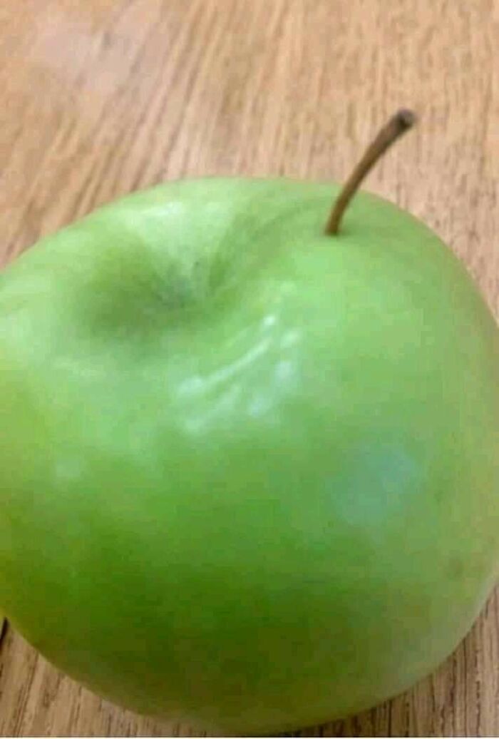Close-up green apple with dent on wooden table, oddity hinting at glitches in the Matrix