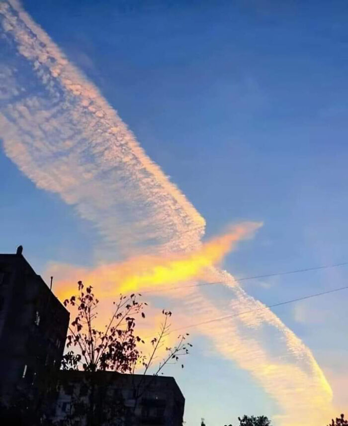Contrail forming X-shaped glow over city skyline at sunset, clouds resembling glitches in the matrix
