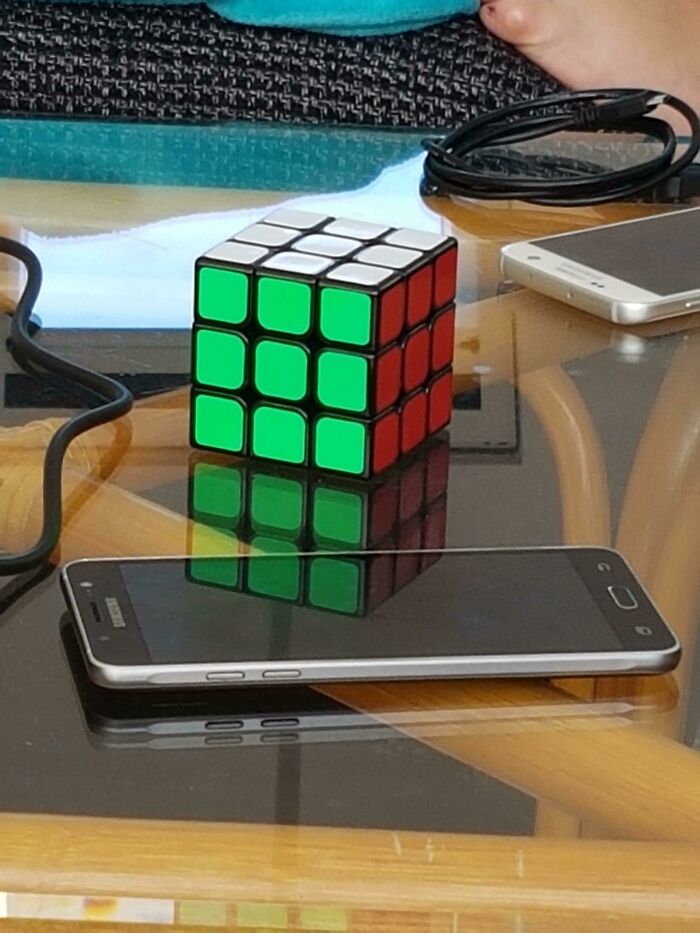 Rubik's Cube with green and red faces reflected on glass table beside smartphones and cords, glitches in the matrix theme