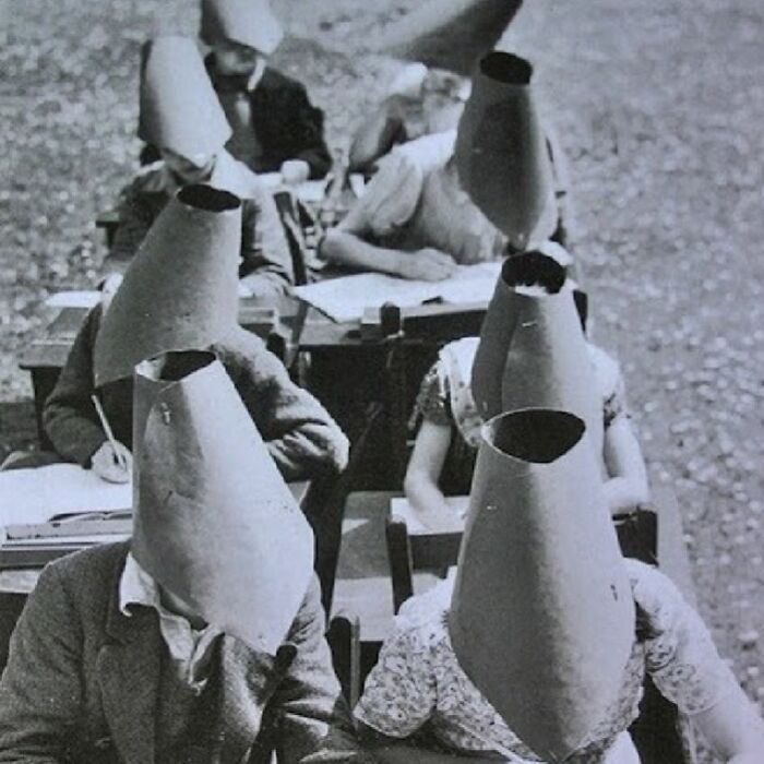 Students wearing tall cone-shaped paper masks while sitting at desks in an old black and white odd and creepy photo.