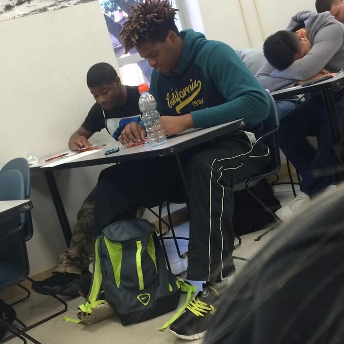 Two tall students sitting at desks in a classroom, one wearing a blue hoodie and struggling with legroom.