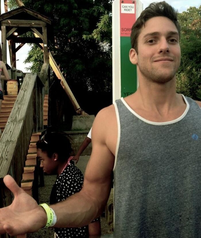 A tall young man in a gray tank top at an outdoor amusement park, illustrating struggles only tall people face.