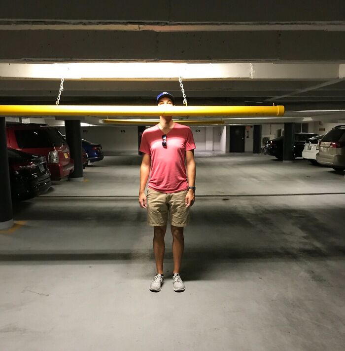 A tall man in a parking garage standing under a yellow barrier illustrating struggles only tall people face.