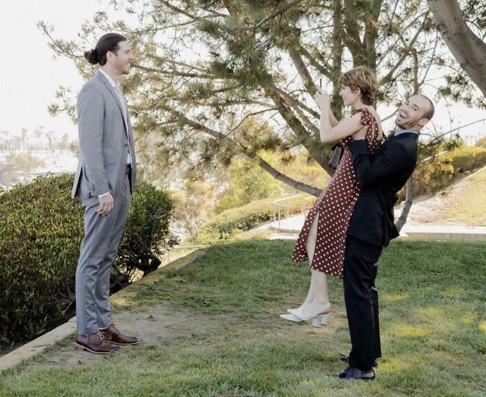 Tall man in a gray suit standing on grass facing a shorter man lifting a woman in a red polka dot dress outdoors.